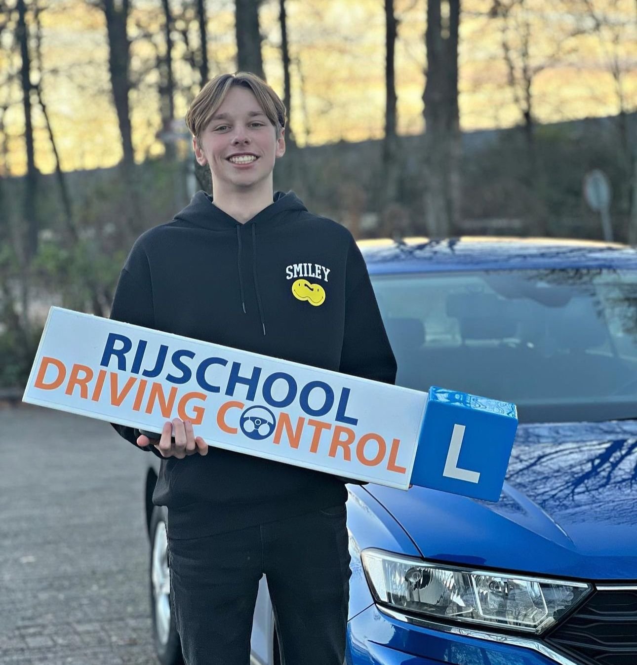 Geslaagde jongen met Rijschool Driving Control bord naast blauwe lesauto — 1 keer geslaagd voor autorijbewijs B automaat in Rotterdam